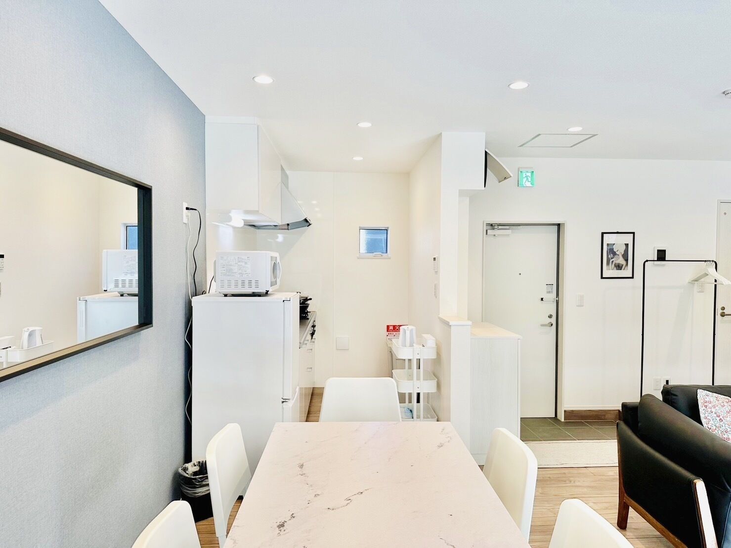 A modern hotel room with a marble-topped dining table, white chairs, a black leather sofa, a microwave, a small refrigerator, and a large mirror on the wall.