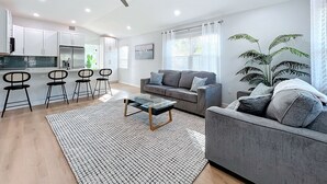 Un salon moderne avec un canapé gris, une table basse et un tapis à motifs. On y trouve également un coin cuisine avec des meubles blancs et un ensemble de salle à manger.