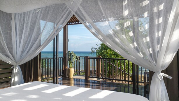 Quarto com uma cama grande, cortinas leves e vista para o oceano.