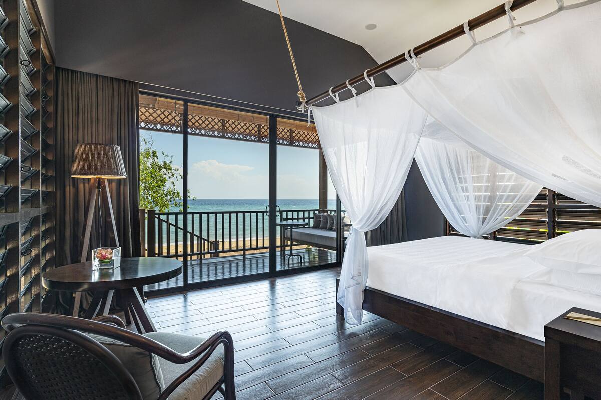 Un dormitorio con cama, una mesa y una silla, un balcón con vista al mar y una silla colgante.