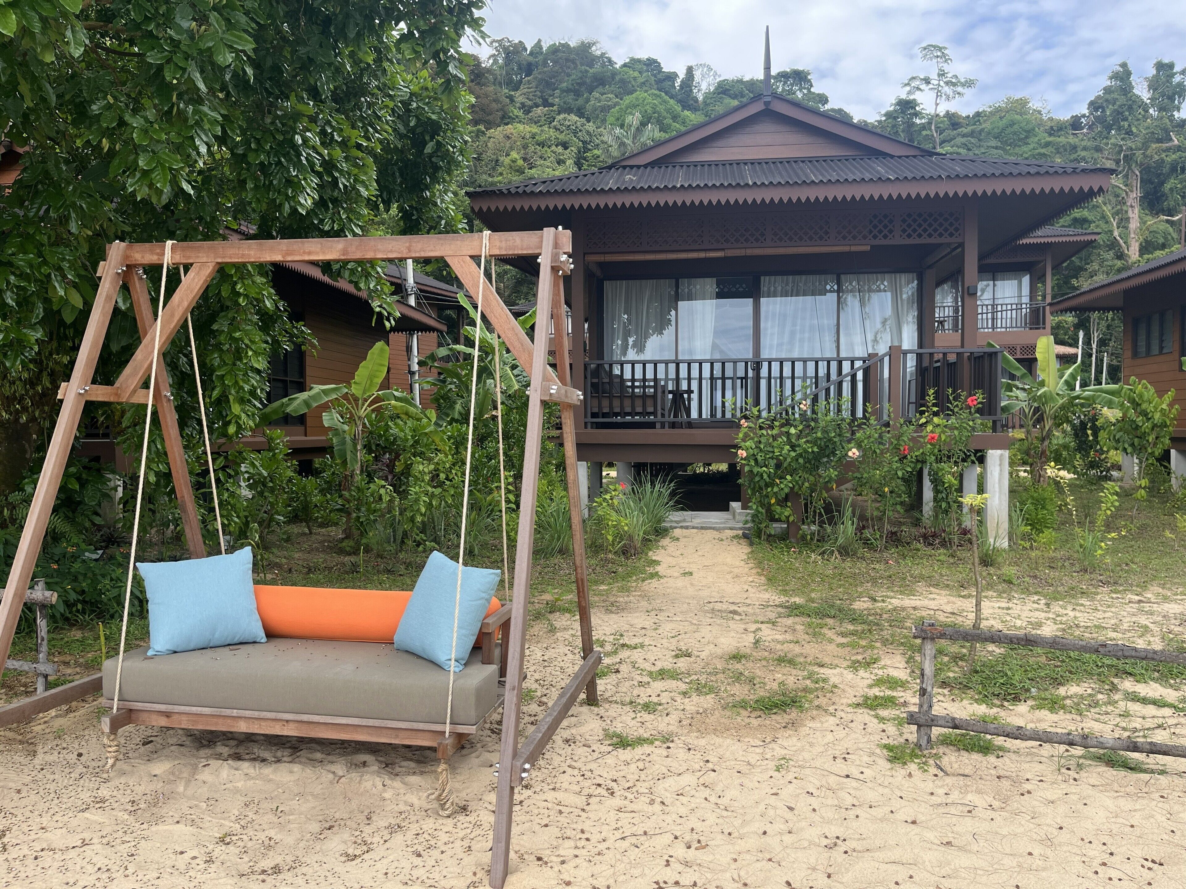 A wooden swing with cushions, a pathway leading to a building with a balcony, surrounded by greenery.