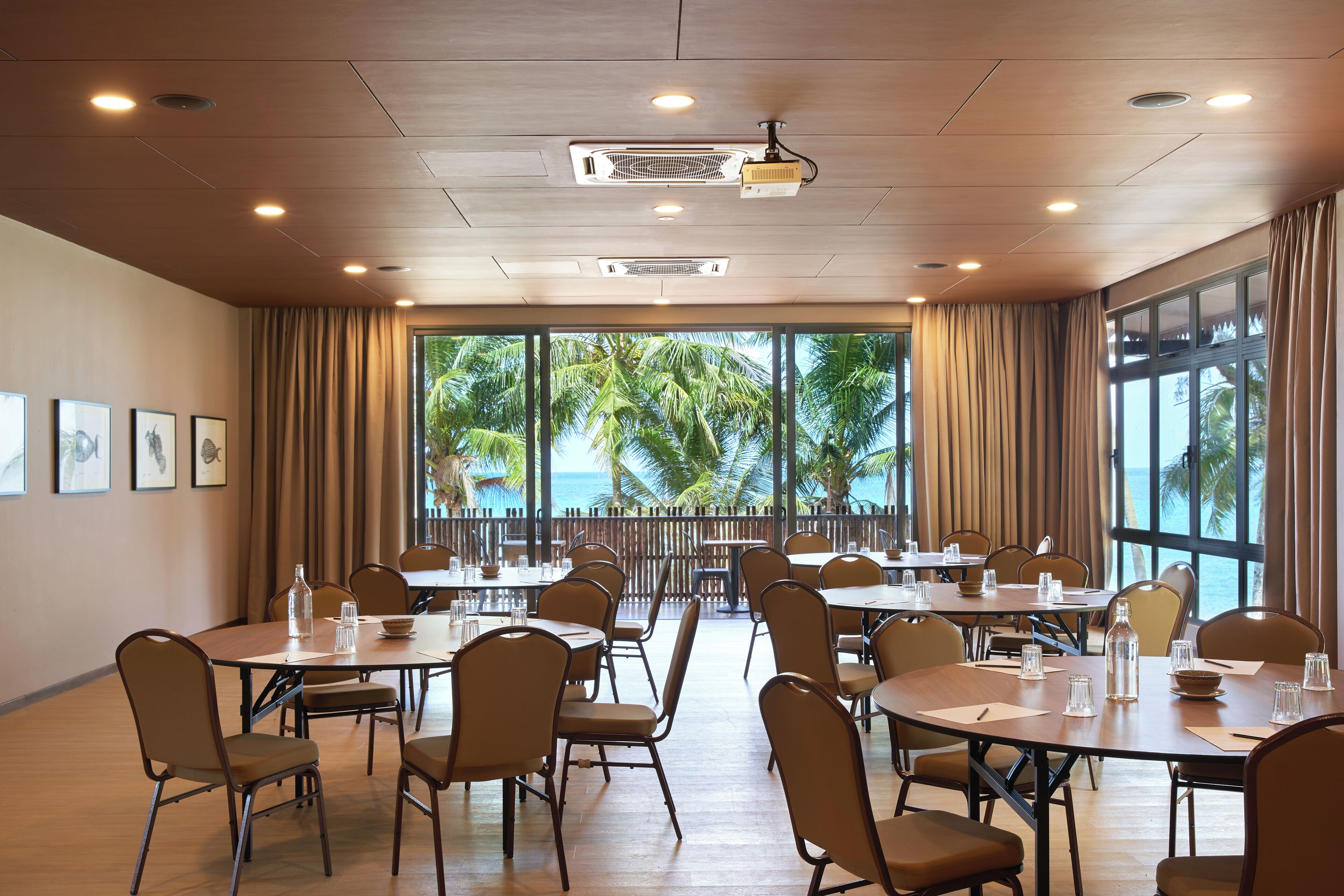 Un salón de conferencias con varias mesas y sillas, ventanas amplias con cortinas e iluminación en el techo.