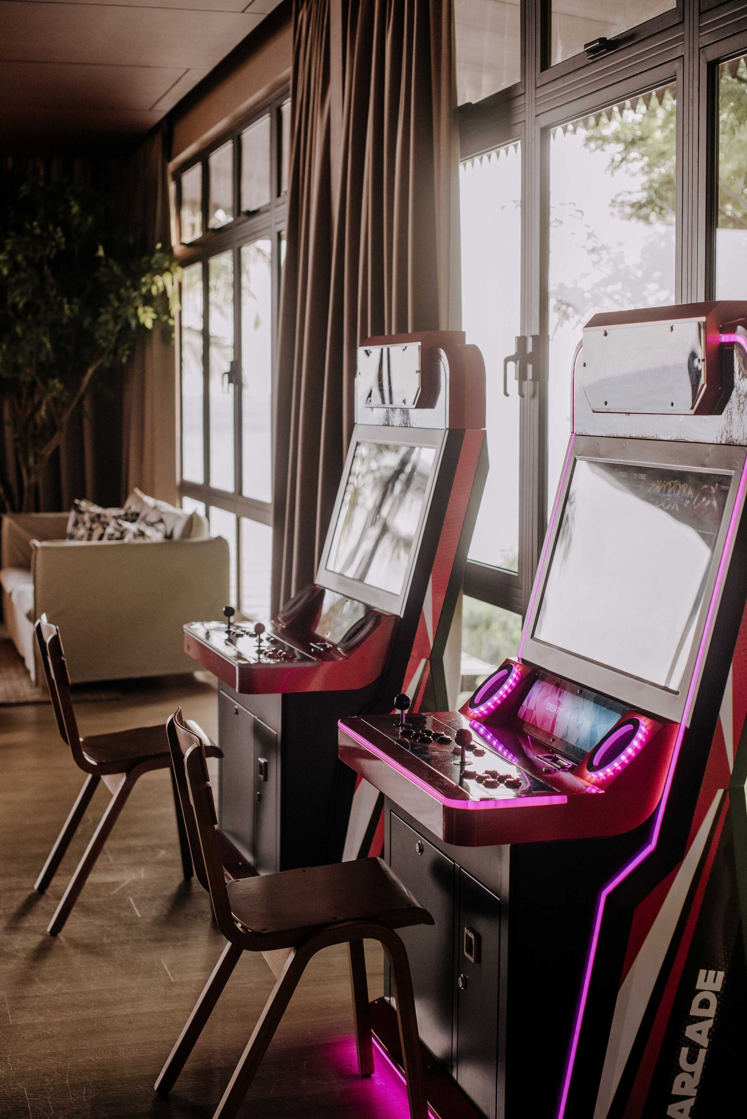 Two arcade machines with neon lights, a sofa, and a window with curtains.