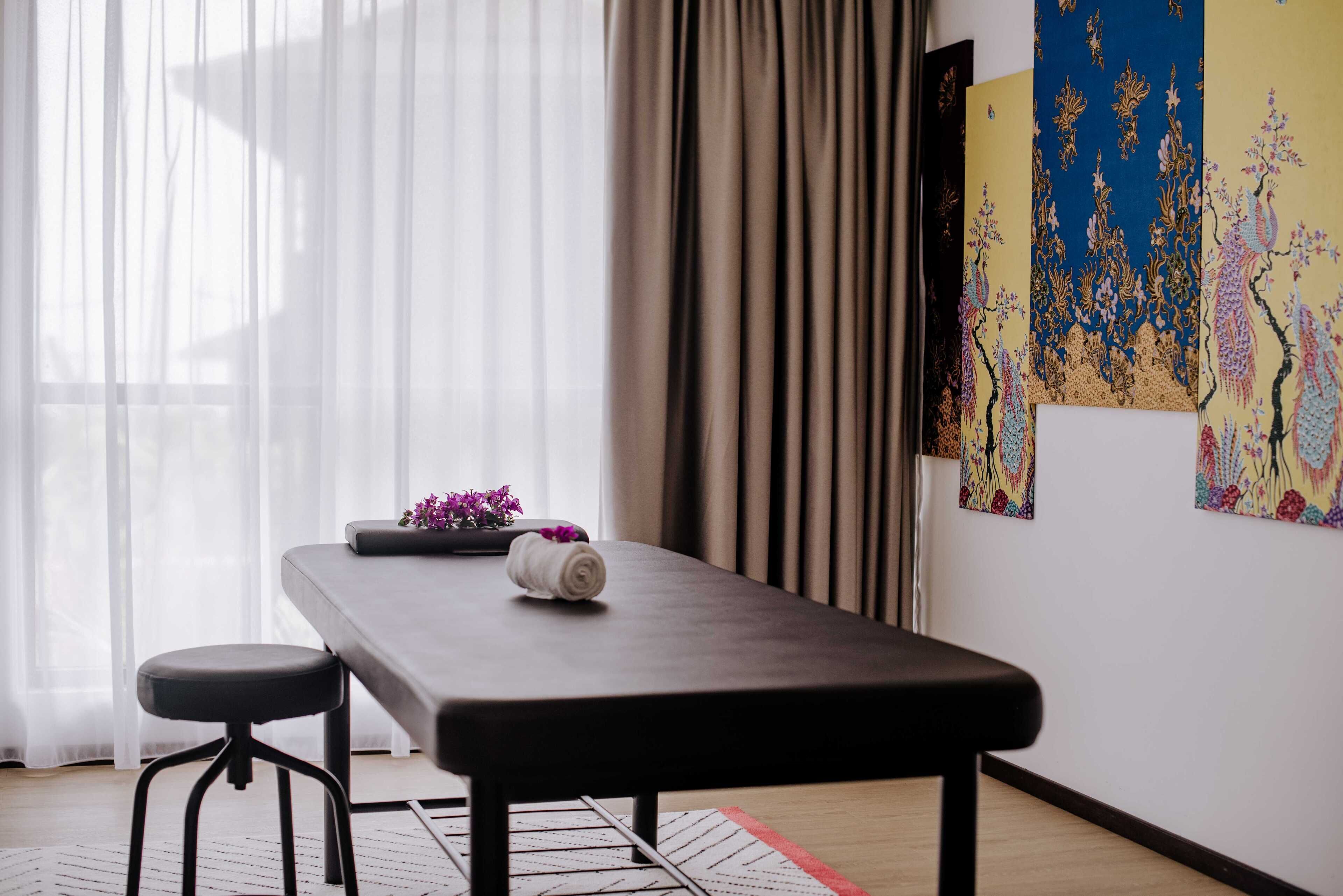 Een kamer met een massagetafel, een klein krukje, een potplant en kunst aan de muur.