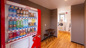 A vending machine with various drinks and snacks, a small table with chairs, and a washing machine in a room with wooden flooring. - HOTEL R9 The Yard Fujioka (Fujioka)