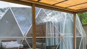 Uma área de estar coberta ao ar livre com um sofá e uma mesa de madeira, uma estrutura de estufa ao fundo e vista para um jardim.