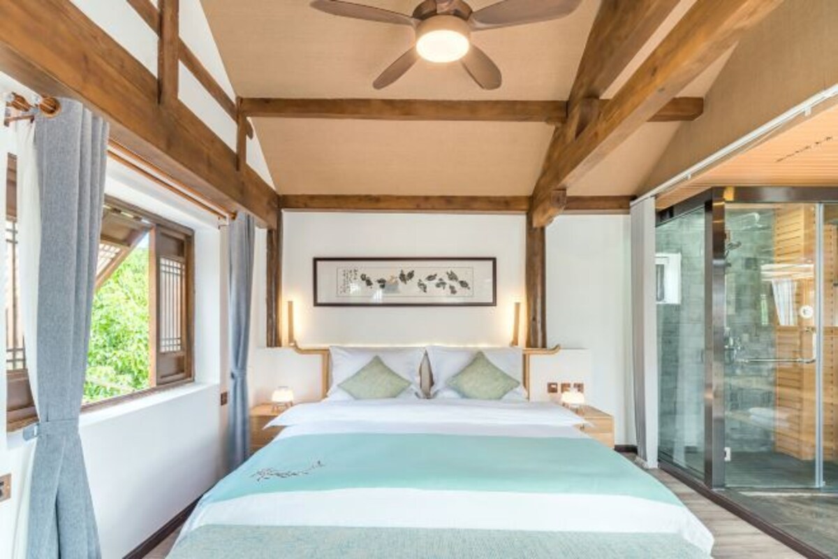A bedroom with a bed, a ceiling fan, a framed picture on the wall, and a glass door leading to a bathroom.