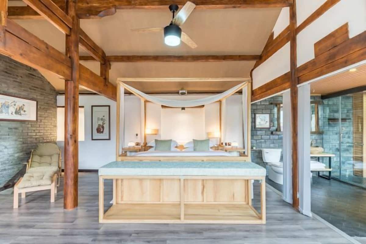 A spacious bedroom with a large bed, wooden beams, a ceiling fan, and a stone wall.