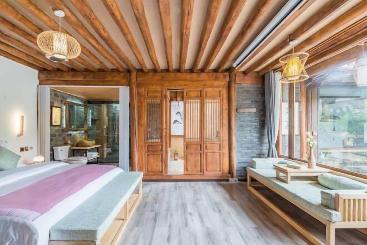 A modern bedroom with a wooden ceiling, a large bed, a bench, and a view of the outdoors.