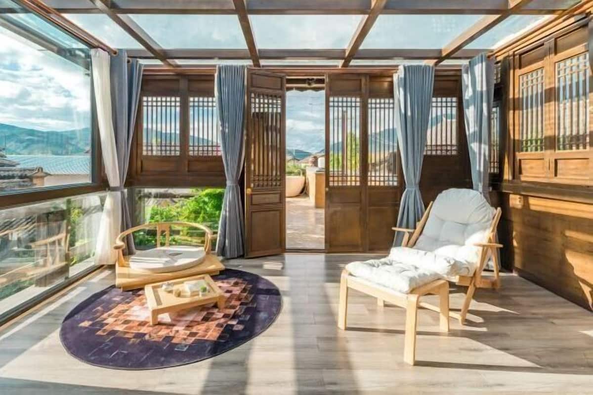 A room with a view of mountains, a wooden chair, a round table, and a large window with curtains.