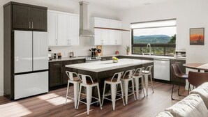 Une cuisine moderne avec des meubles blancs, un îlot central doté de tabourets, une grande fenêtre offrant une belle vue et un coin repas avec une table et des chaises.
