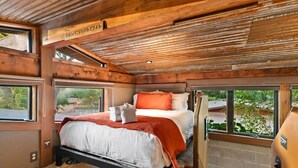 Une chambre avec un plafond en bois, un lit avec une literie blanche et un oreiller orange, et une vue sur la verdure à l’extérieur.