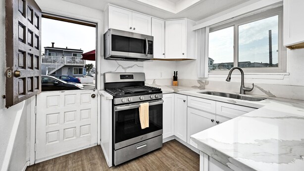 A modern kitchen with white cabinetry, a stainless steel stove, a microwave, and a large window providing a view of the neighborhood. - Charming 1-Bedroom Home on 29th Street (Newport Beach)