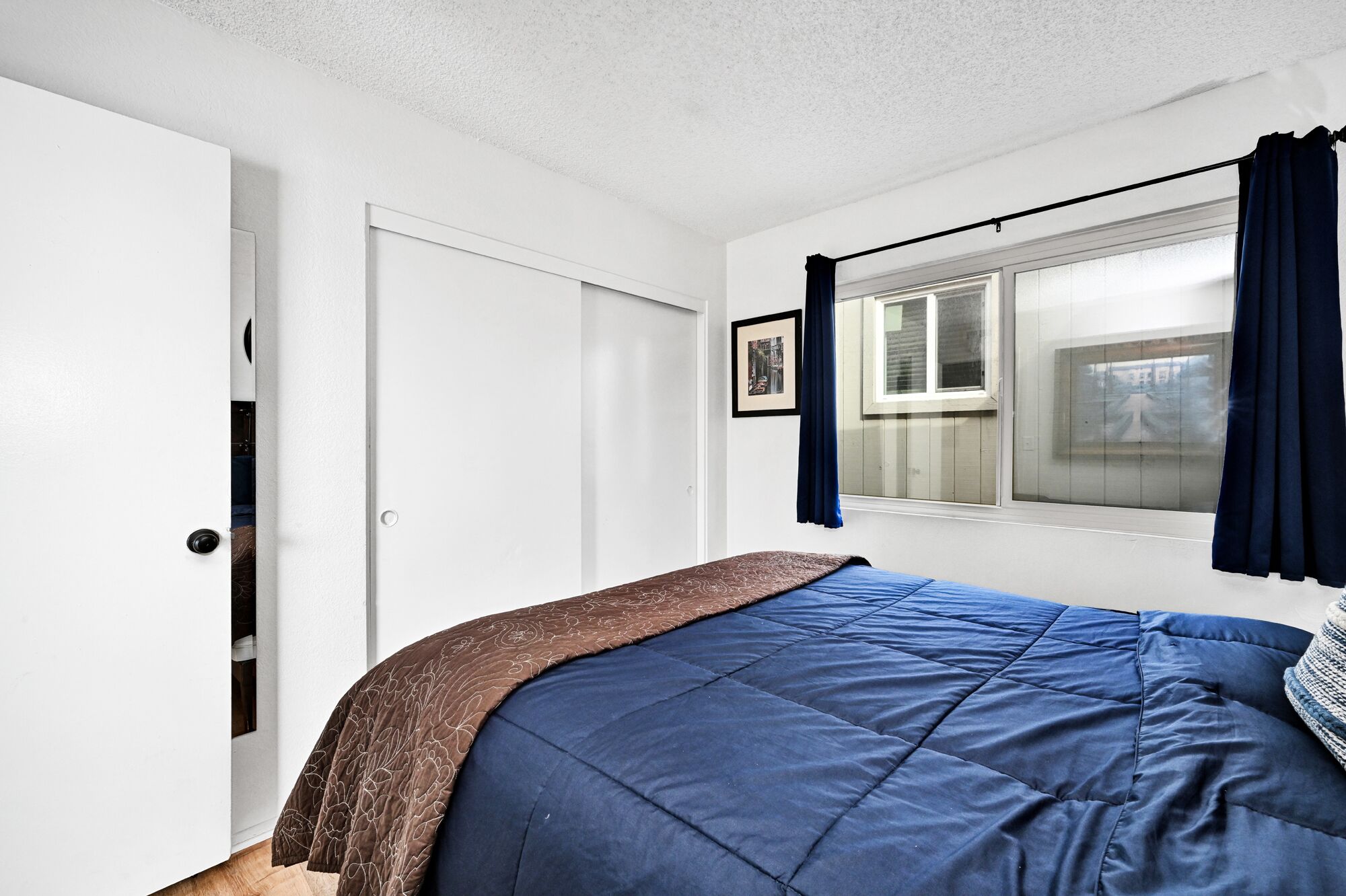 A bedroom with a bed, a window with curtains, a door, and a framed picture on the wall.