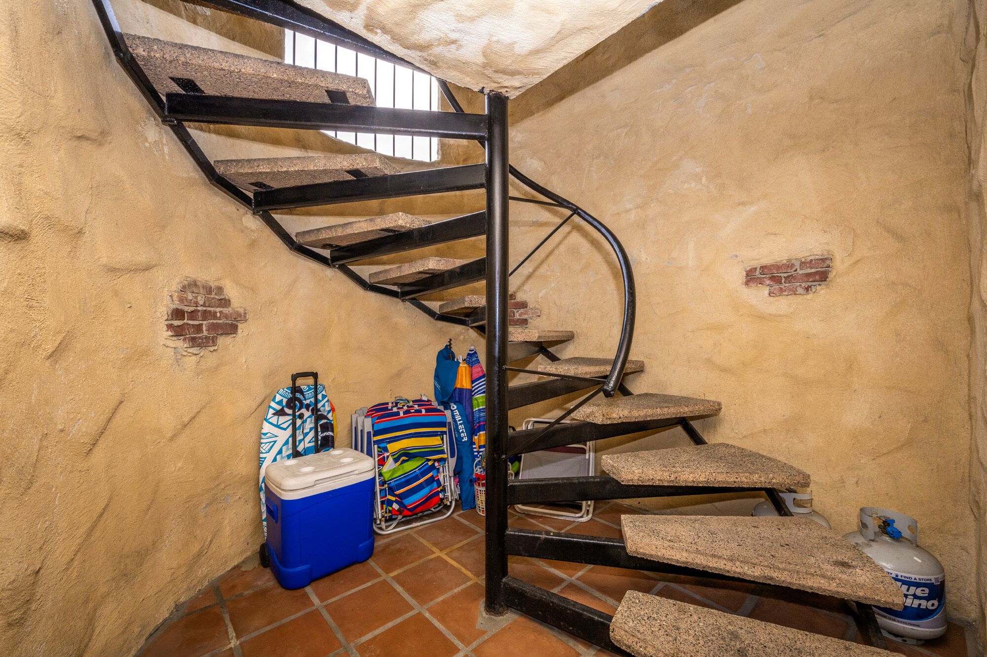 A spiral staircase with stone steps and metal railings, a blue cooler, colorful bags, and a propane tank in a rustic room with brick walls.