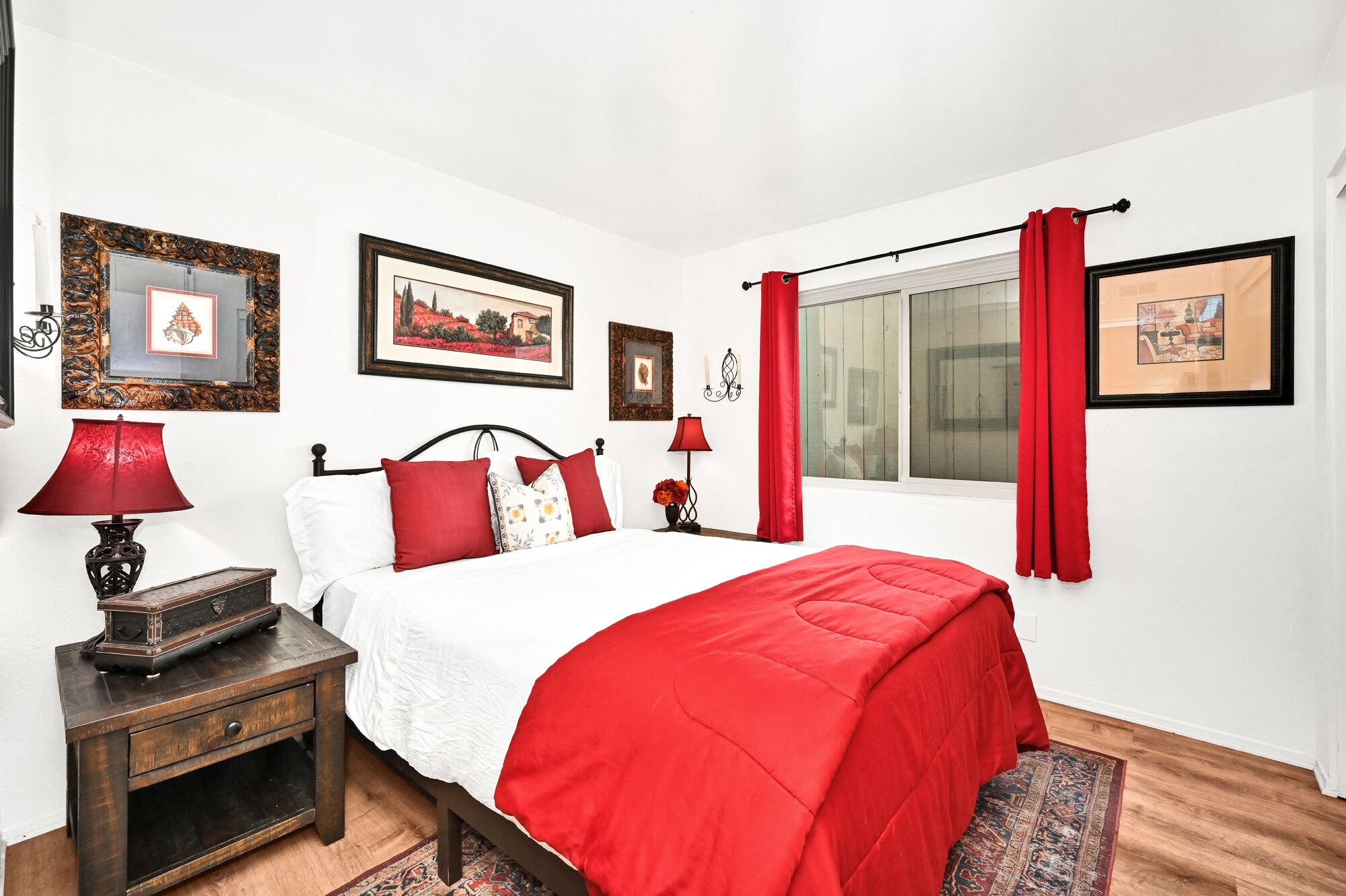 Un dormitorio con una cama, una mesita de noche, una lámpara y una ventana con cortinas rojas.