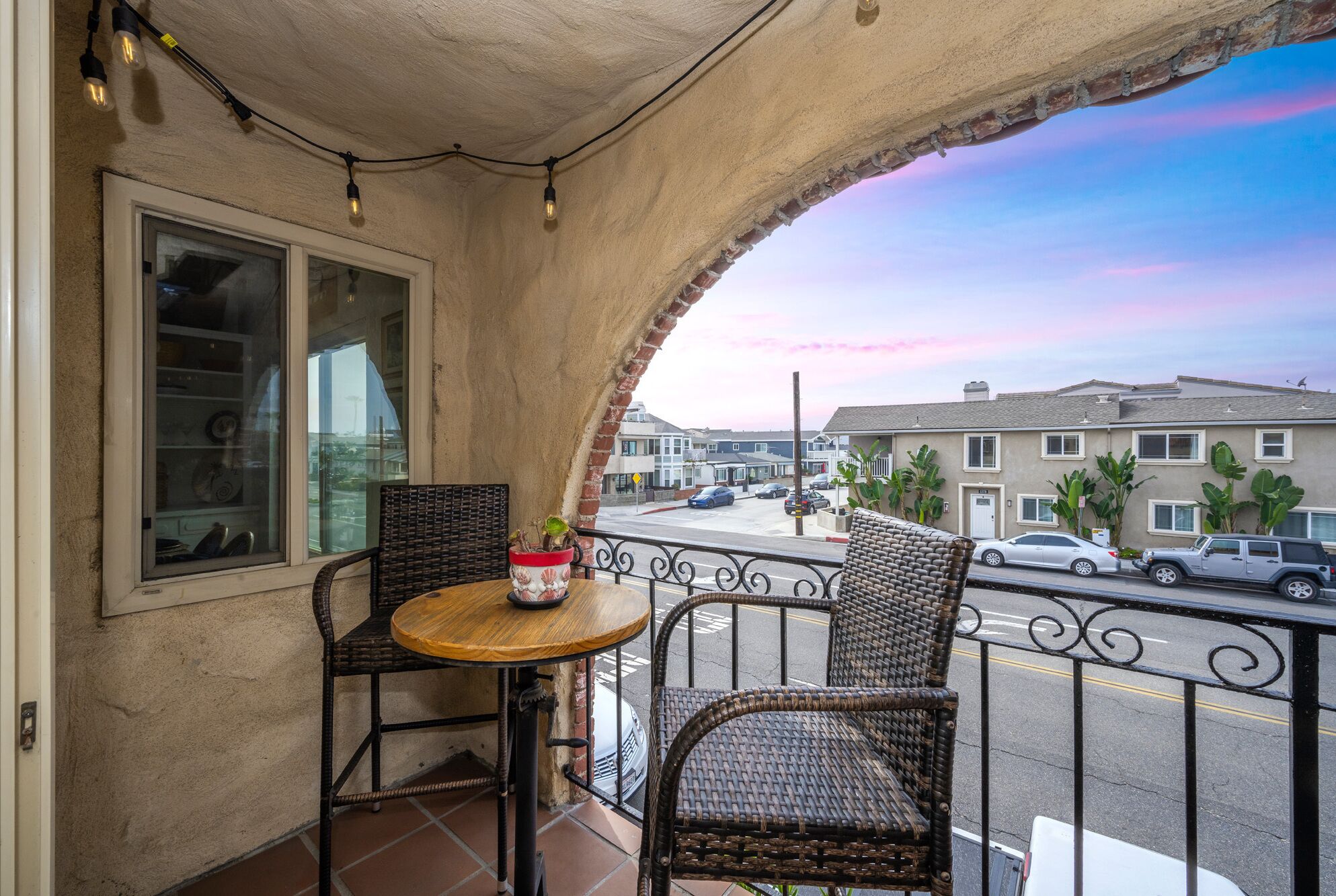 A balcony with wicker chairs and a small table, overlooking a street with parked cars and buildings.