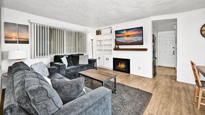 Un salon moderne avec un canapé gris, une table basse et une cheminée. Un téléviseur est fixé au mur et il y a une fenêtre avec des stores.