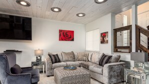 A living room with a sectional sofa, ottoman, and a TV mounted on the wall. - Lovely 2BA King Beds+2 Car Garage. Hidden Gem-Walk>Shops & Dining! (Littleton)