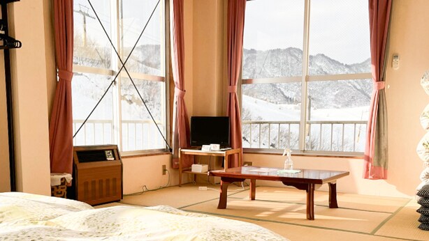 A hotel room with a bed, a desk with a chair, a television, and a view of snowy mountains through large windows. - ishiuchiskicenter (Minamiuonuma)
