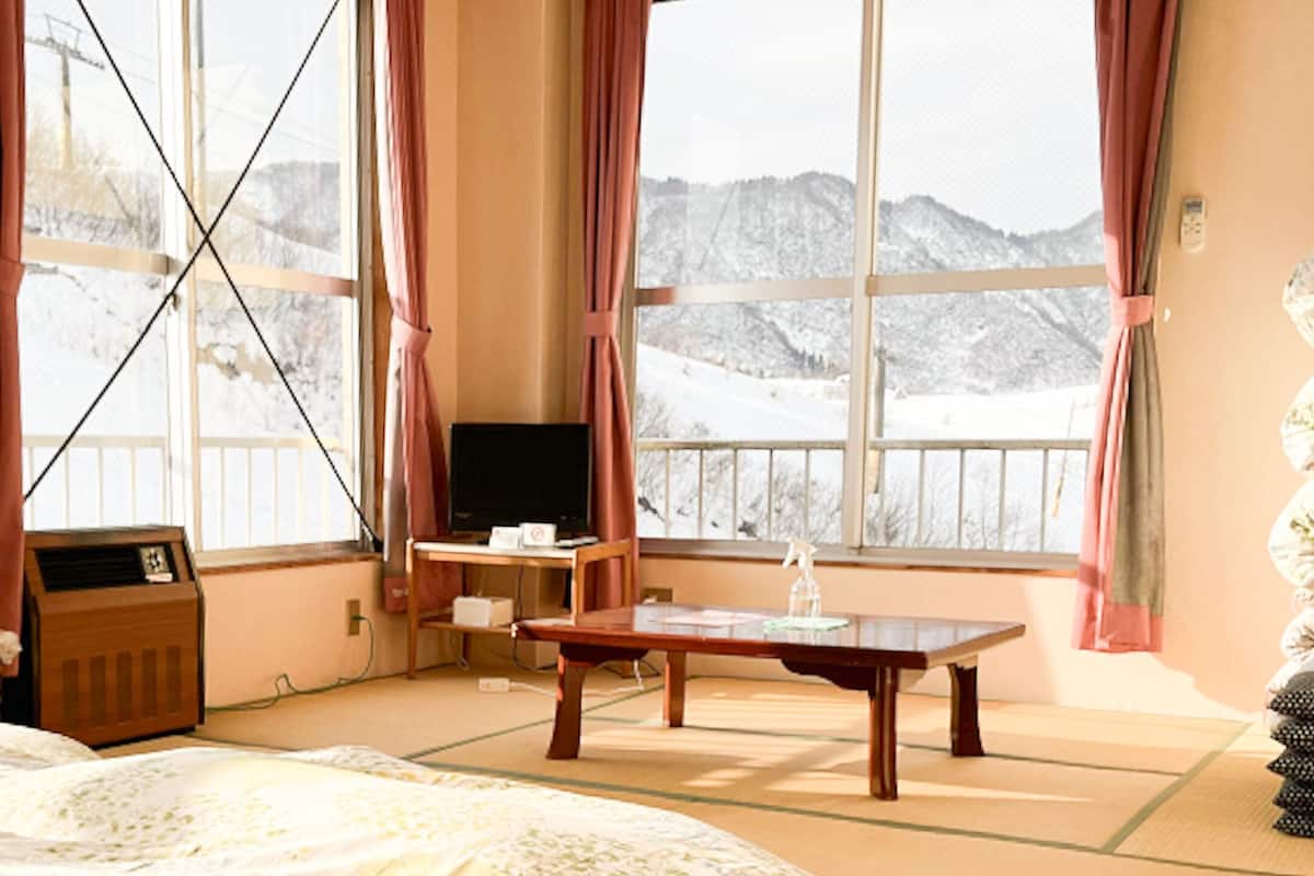 Ein Hotelzimmer mit einem Bett, einem Schreibtisch mit Stuhl, einem Fernseher und Blick auf verschneite Berge durch große Fenster.