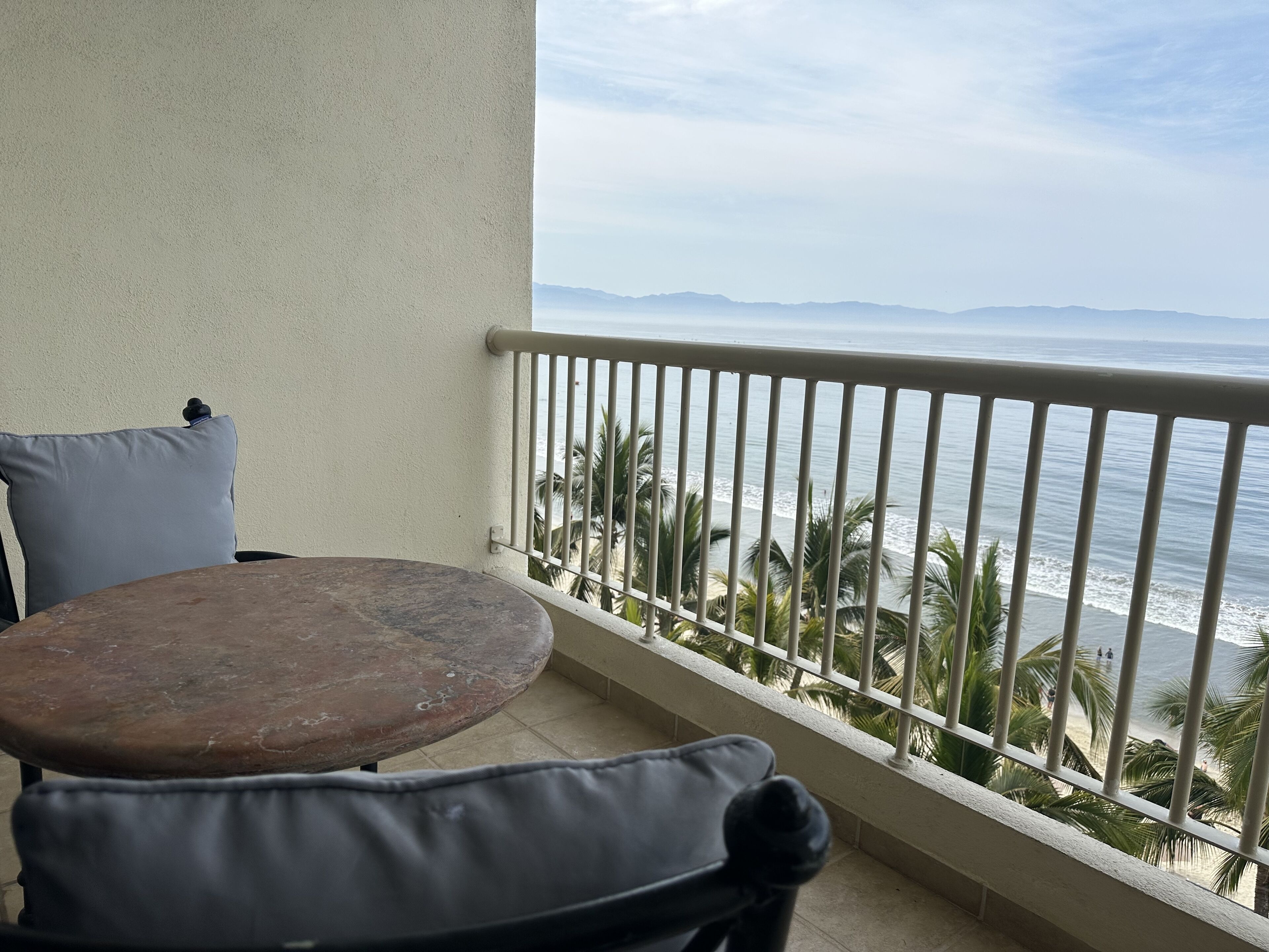 A balcony with a table and chairs, overlooking a beach with palm trees and the ocean.