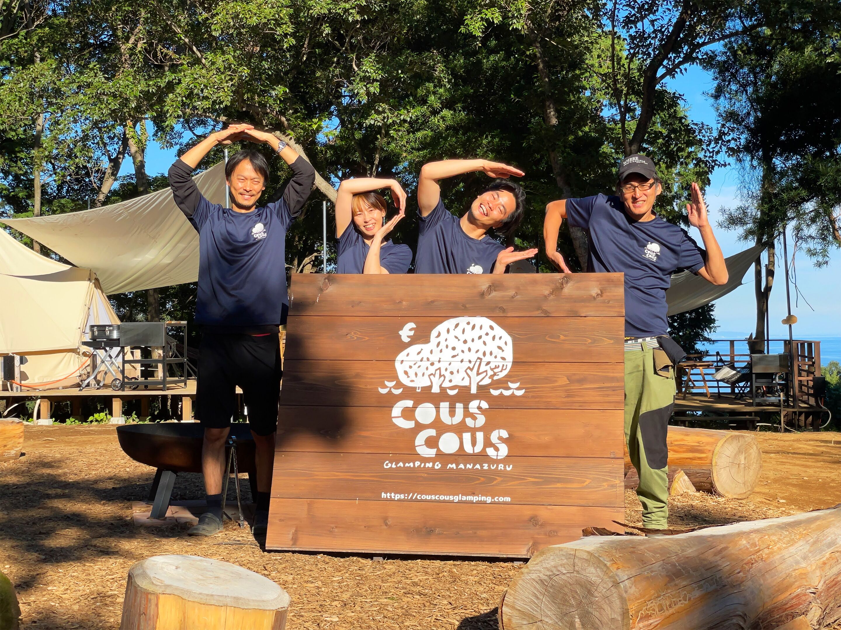 A group of people posing in front of a wooden sign with a tree logo and text, set in an outdoor area with a tent and camping equipment.