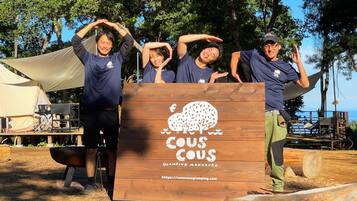 A group of people posing in front of a wooden sign with a tree logo and text, set in an outdoor area with a tent and camping equipment.