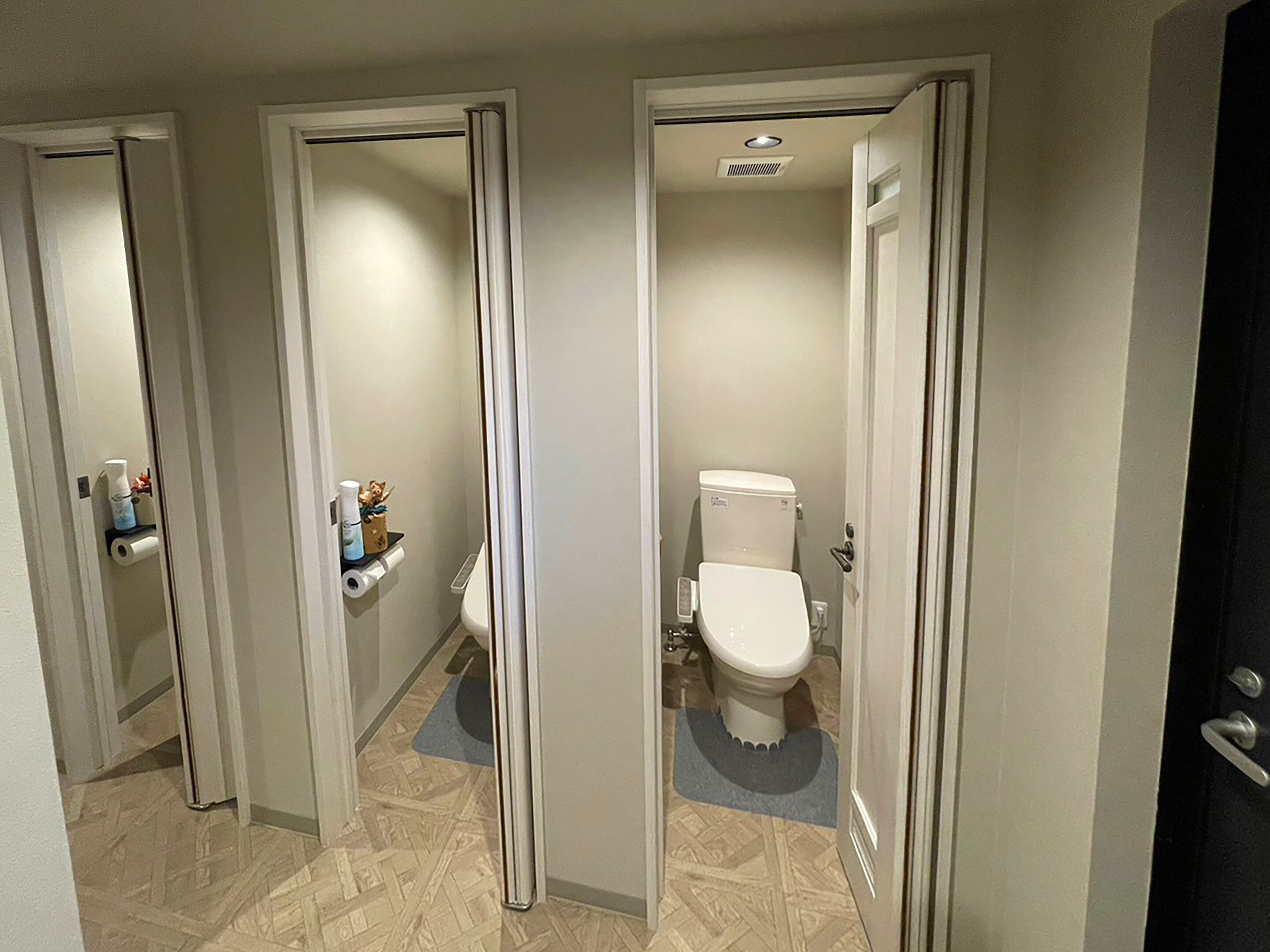 Ein Flur mit drei zur Toilette führenden Türen. Jedes Badezimmer verfügt über ein WC und ein Waschbecken. Der Boden ist mit Fliesen ausgelegt, und im Flur liegt eine Fußmatte in der Mitte.