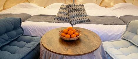 A cozy tent with a bed, a round wooden table with a basket of oranges, and blue and gray cushions.