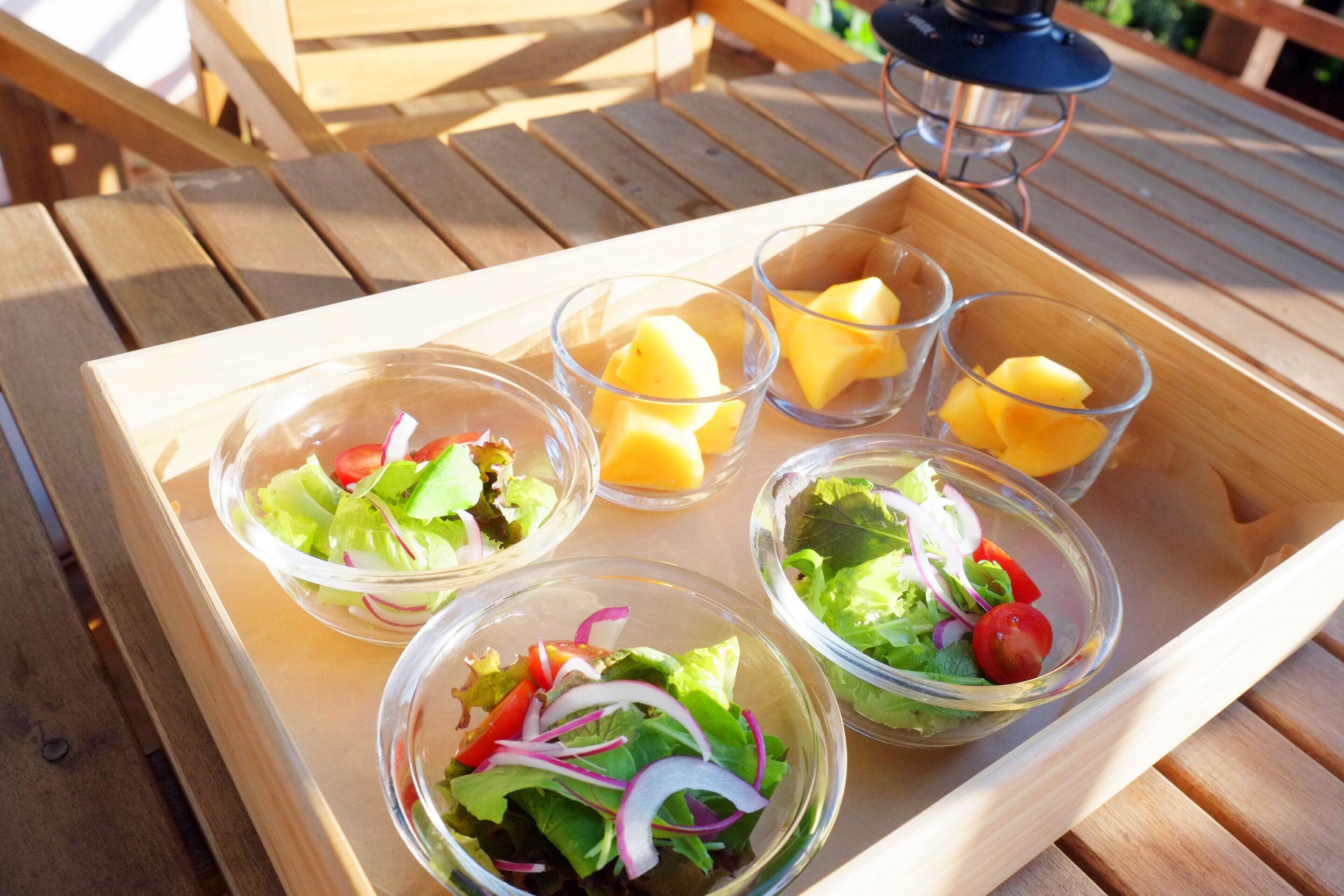 Una mesa de madera con una bandeja que sostiene cuatro recipientes de vidrio con ensaladas y trozos de mango.