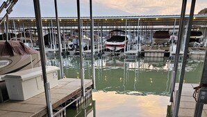Ein Yachthafen mit vertäuten Booten, ein überdachter Bereich mit Bänken und eine ruhige Wasseroberfläche, die den Himmel spiegelt.