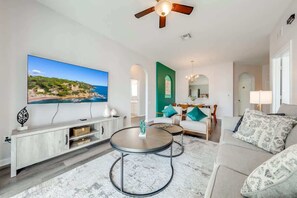 Un salon doté d’un grand téléviseur à écran plat, d’un ventilateur de plafond, d’un canapé, d’une table basse et d’un coin repas avec une table et des chaises.
