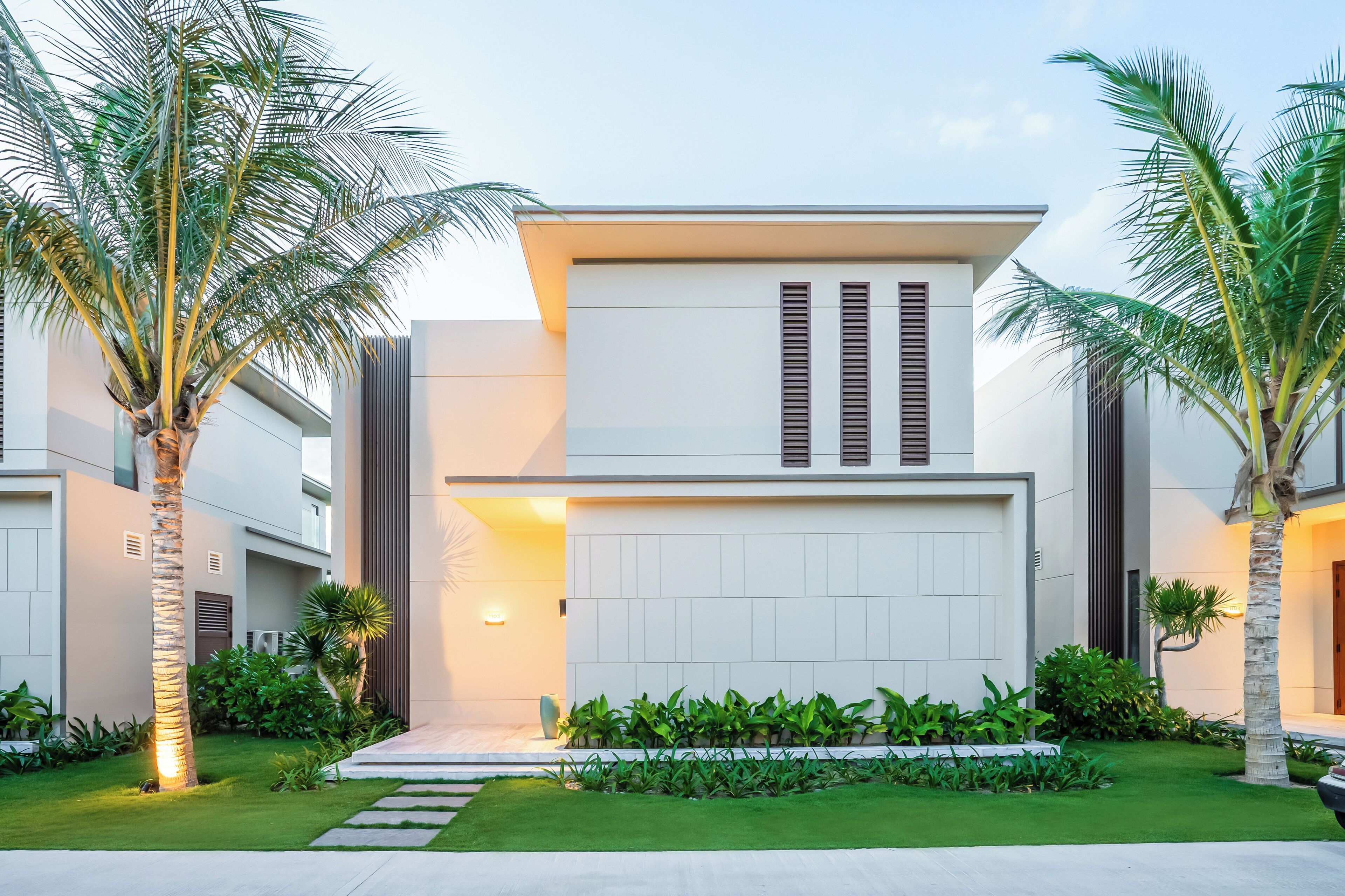 Una moderna casa a due piani con facciata bianca, un ampio garage e un giardino curato con palme e arbusti.