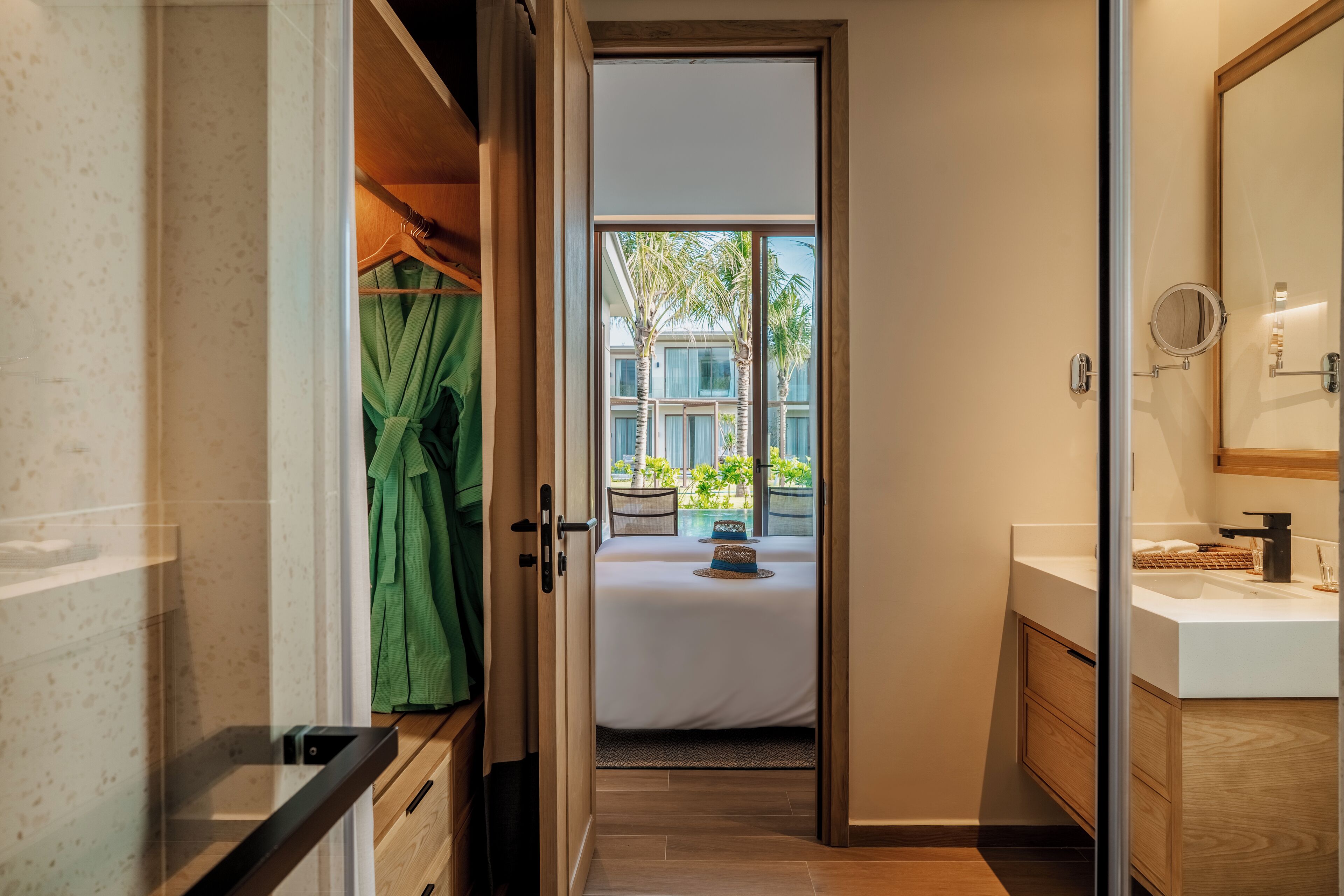 A hotel room with a bed, a bathroom with a mirror and sink, and a view of palm trees and buildings outside.