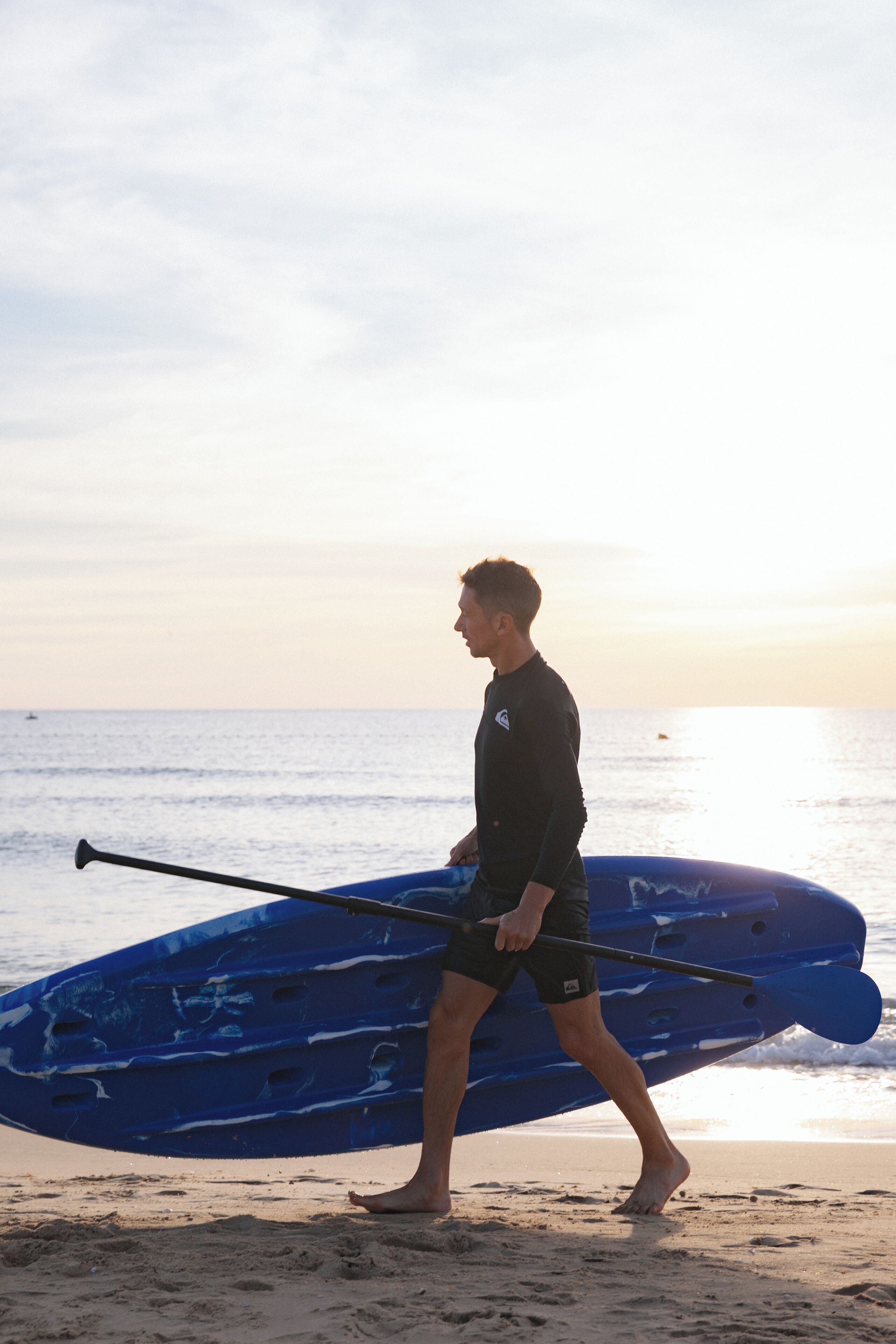 Une personne transportant une planche de surf bleue et une pagaie sur une plage.
