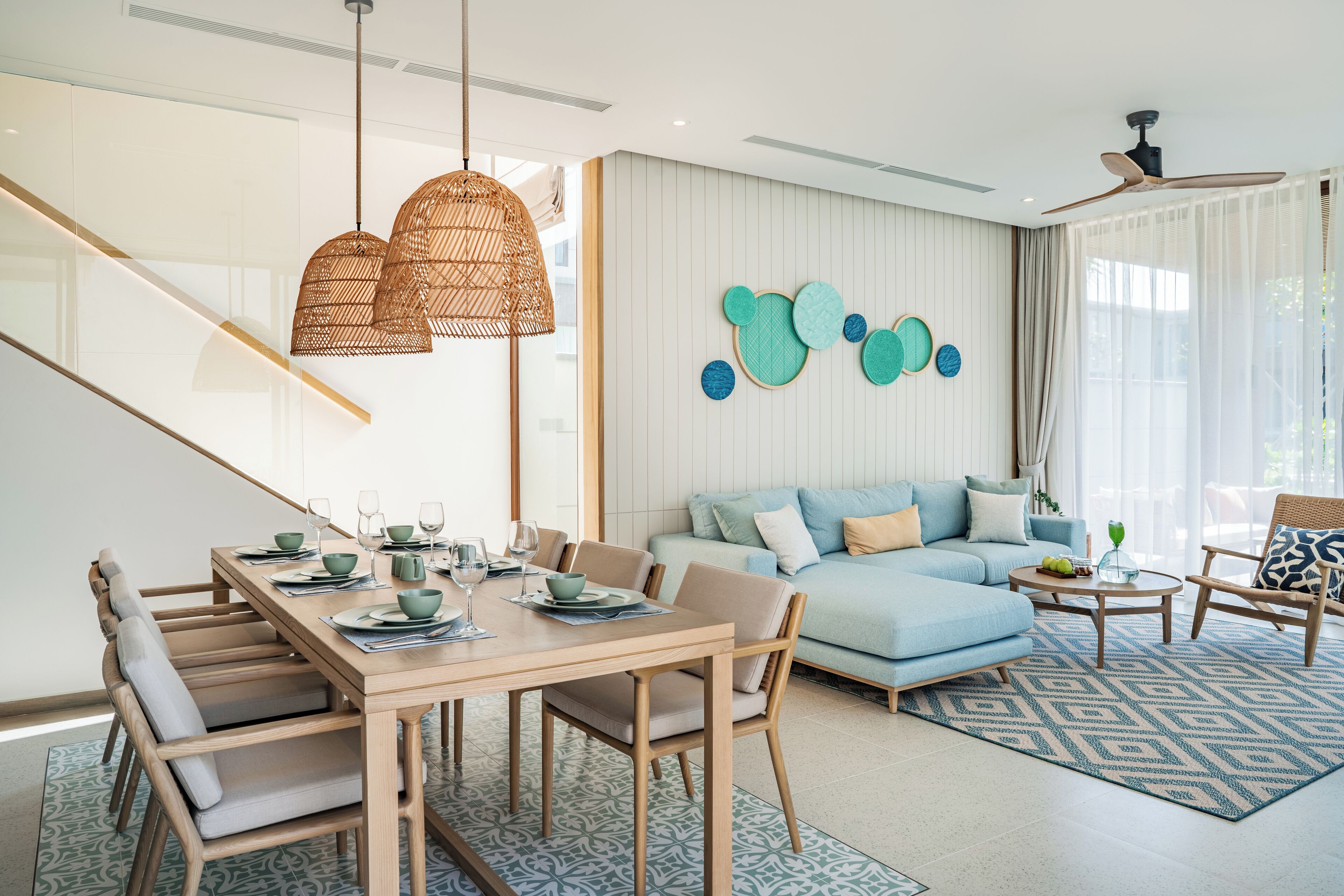 A modern living room with a dining area, featuring a staircase, a blue sofa, and a ceiling fan.