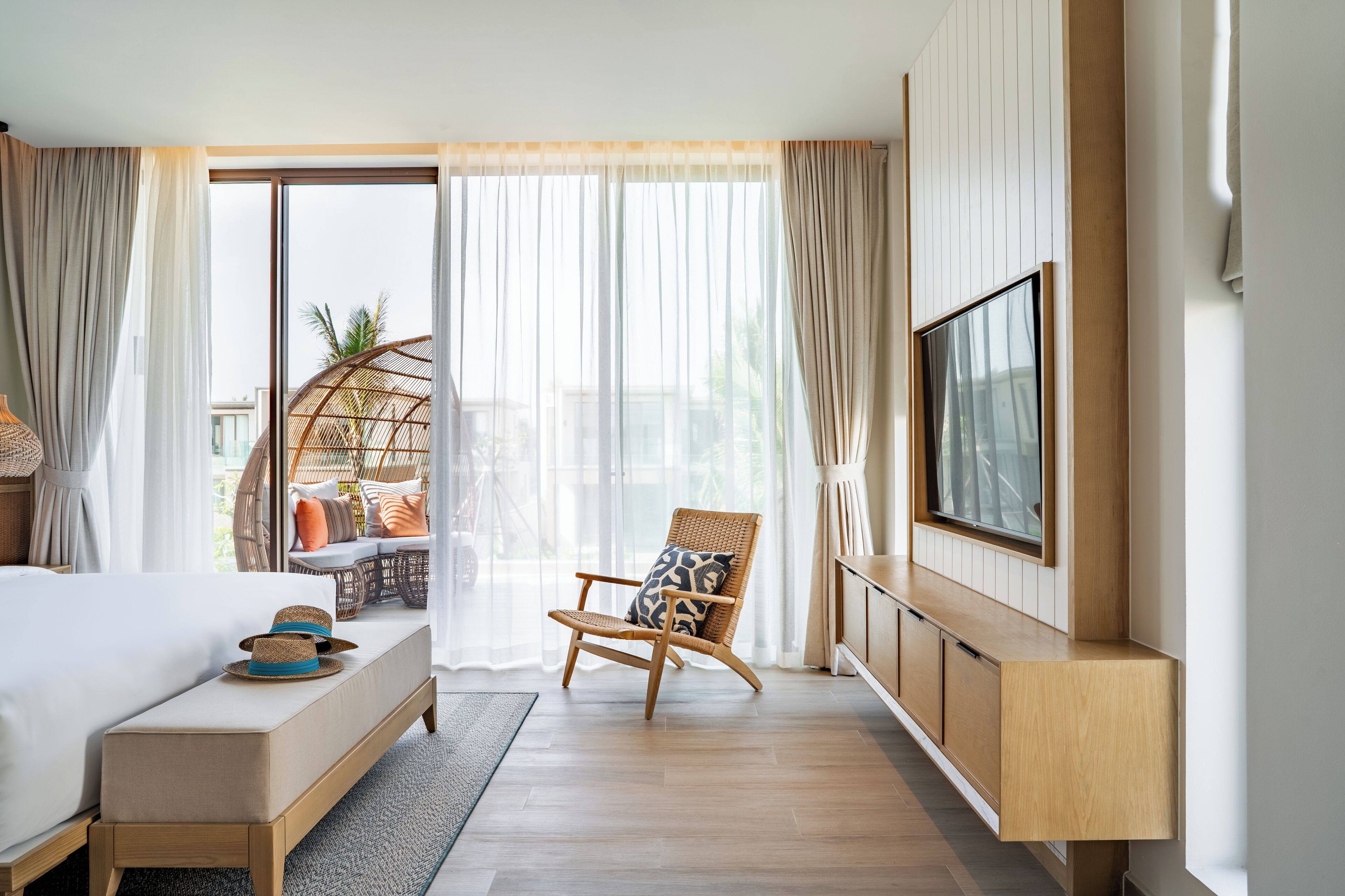 Et moderne hotelværelse med en stor seng, en træbænk, en fletstol, et tv ophængt på væggen og en skydedør af glas, der fører til et udendørs siddeområde.