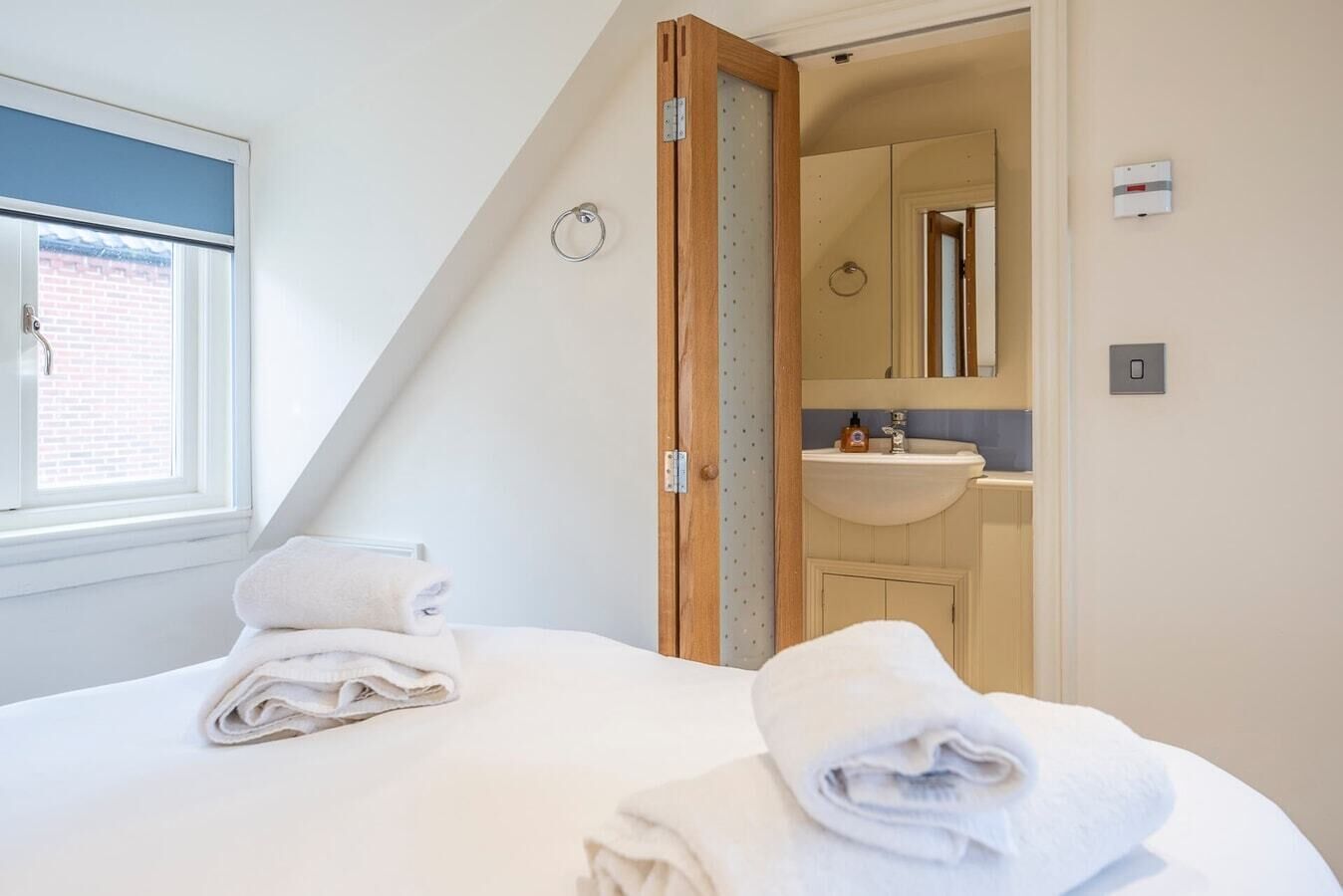 A bedroom with a bed, two towels, a window, and a view of a bathroom with a sink and mirror.