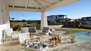 An outdoor seating area with a pool, featuring white furniture and a table set with cups and a fruit bowl.