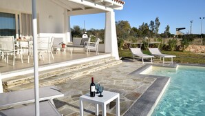 An outdoor pool area with lounge chairs, a table with a bottle of wine, and a pergola structure.
