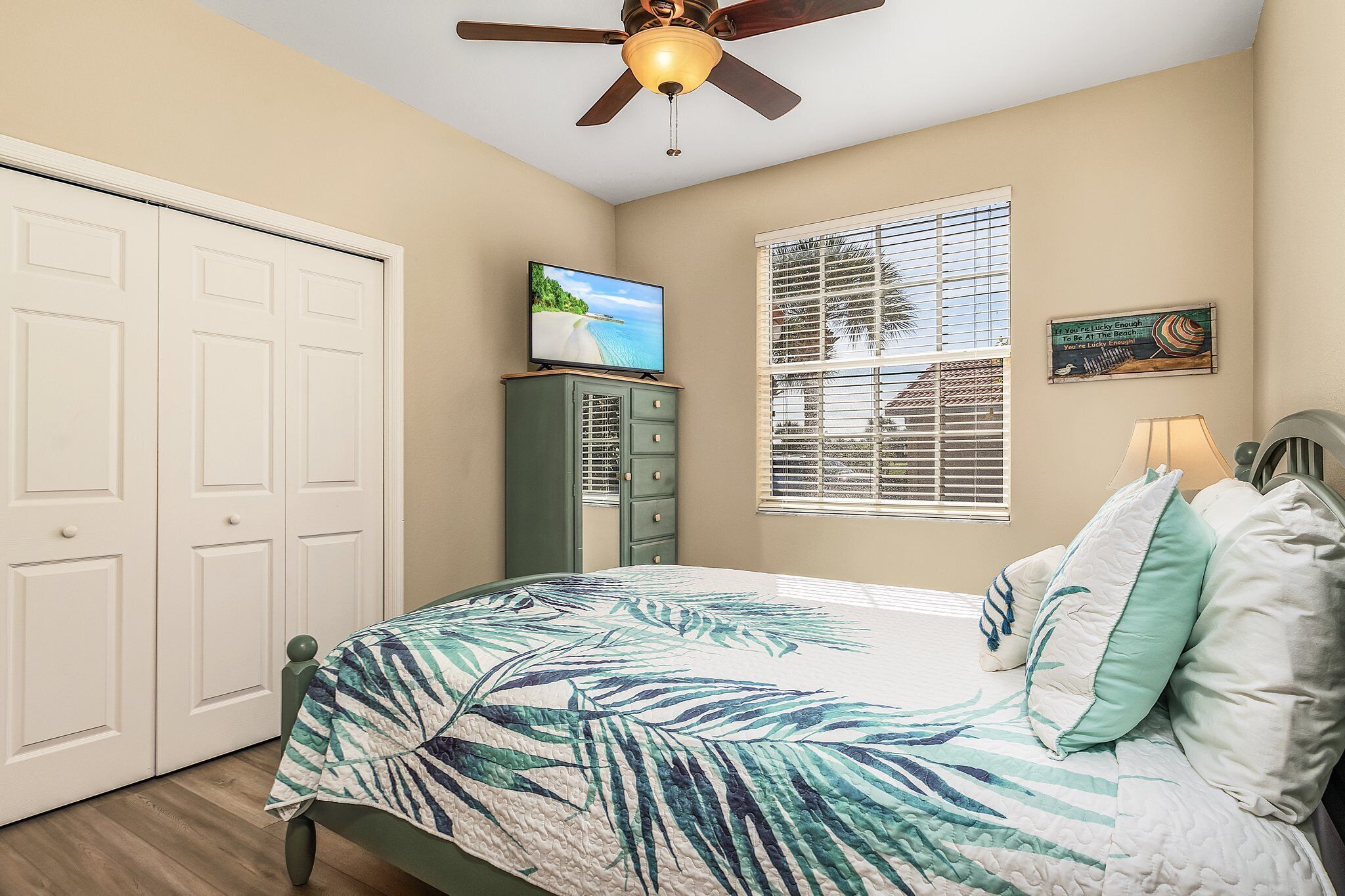 A bedroom with a bed, a ceiling fan, a TV on a dresser, a window with blinds, and a door.