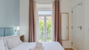 A hotel room with a bed, a nightstand, a lamp, a door, and a balcony with curtains. - Downtown Setúbal Apartment (Setúbal)