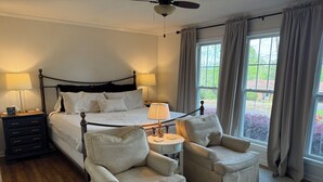 Un dormitorio con una cama, dos mesitas de noche con lámparas, una mesita redonda auxiliar, dos sillones, un ventilador de techo y una ventana con cortinas.