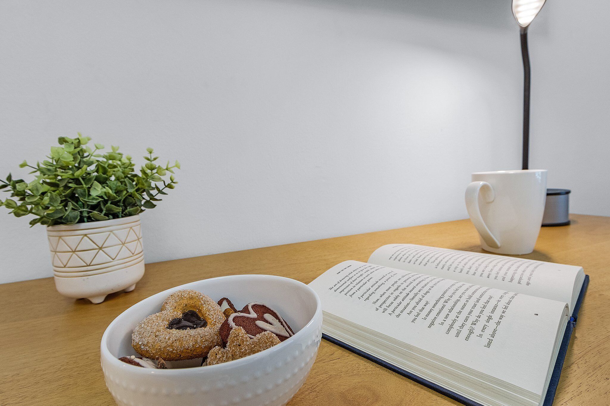 A wooden table with a bowl of pastries, a potted plant, a white mug, and an open book.