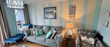A living room with a grey sofa, blue and patterned cushions, a small table, and a beach-themed chair.