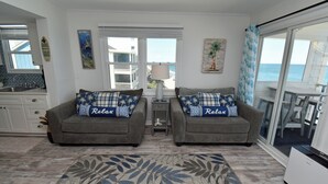 A living room with two grey sofas, a patterned rug, a small table with a lamp, and a view of the ocean through large windows. - Carolina Shores II 1404 - Oceanfront Complex in Garden City Beach (Garden City Beach)