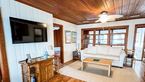 A spacious living room with wooden floors, a white sofa, a coffee table, a TV mounted on the wall, and a ceiling fan. - Inlet Holiday - Creekfront in Garden City Beach (Garden City Beach)