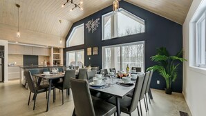 A modern kitchen with dark walls and large windows, a dining area with a table set for six, and a potted plant in the corner. - The Experience SÔ Lodge (SHEFFORD)