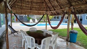 An outdoor dining area with a thatched roof, a pool in the background, and a trash bin to the side.